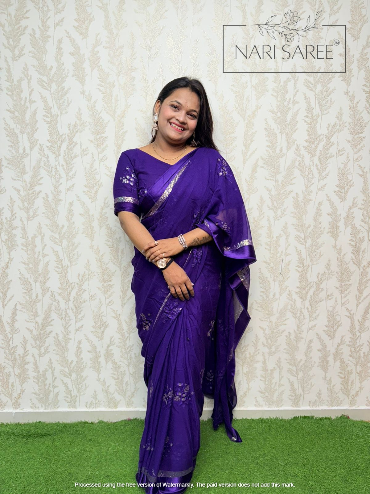 Woman wearing a purple saree with silver border against a textured white wall.